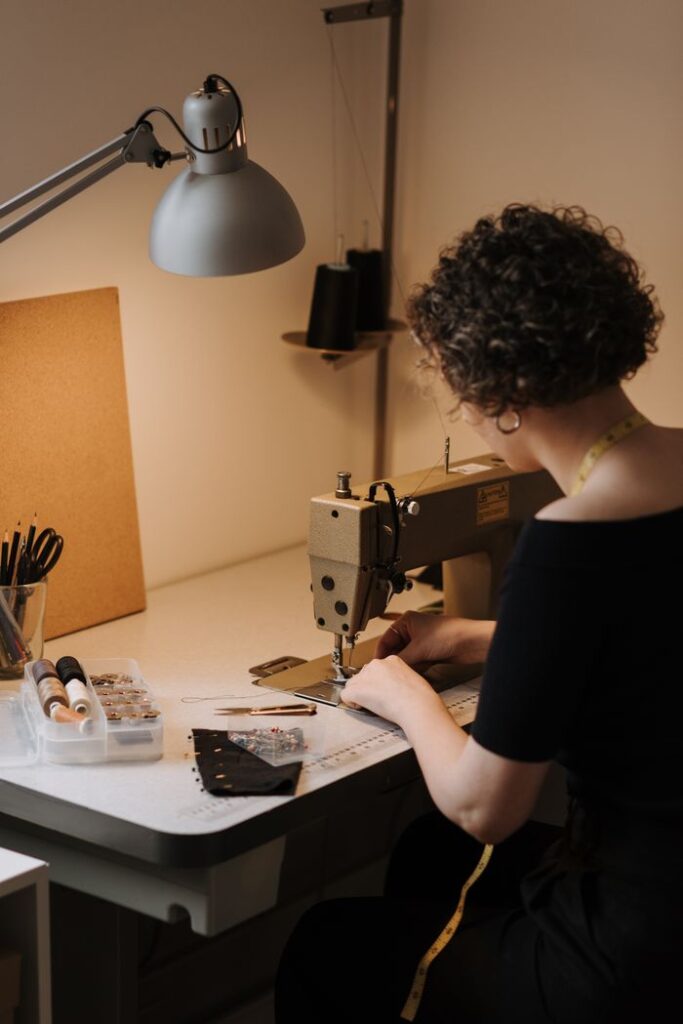 Side view of skilled faceless female dressmaker working on professional sewing machine at table with sewing supplies in modern workshop